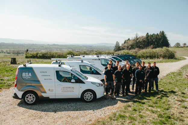 A group of electricians stood in front and to the right of a line of 8 vans.